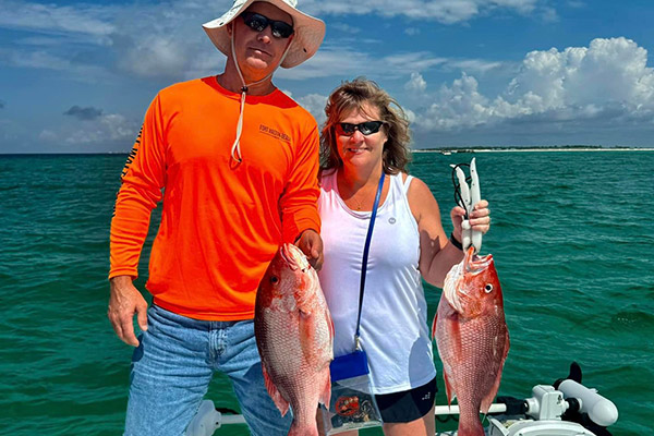 man wearing tan boaters hat with a bright orange shirt holding a red snapper standing next to a woman wearing a white tank top also holding a red snapper
