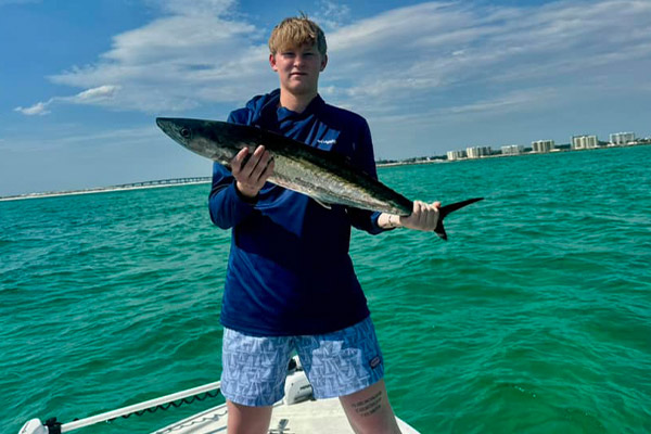 man in a navy blue shirt and blue swim shorts holding a large fish, behind him is the emerald waters of destin